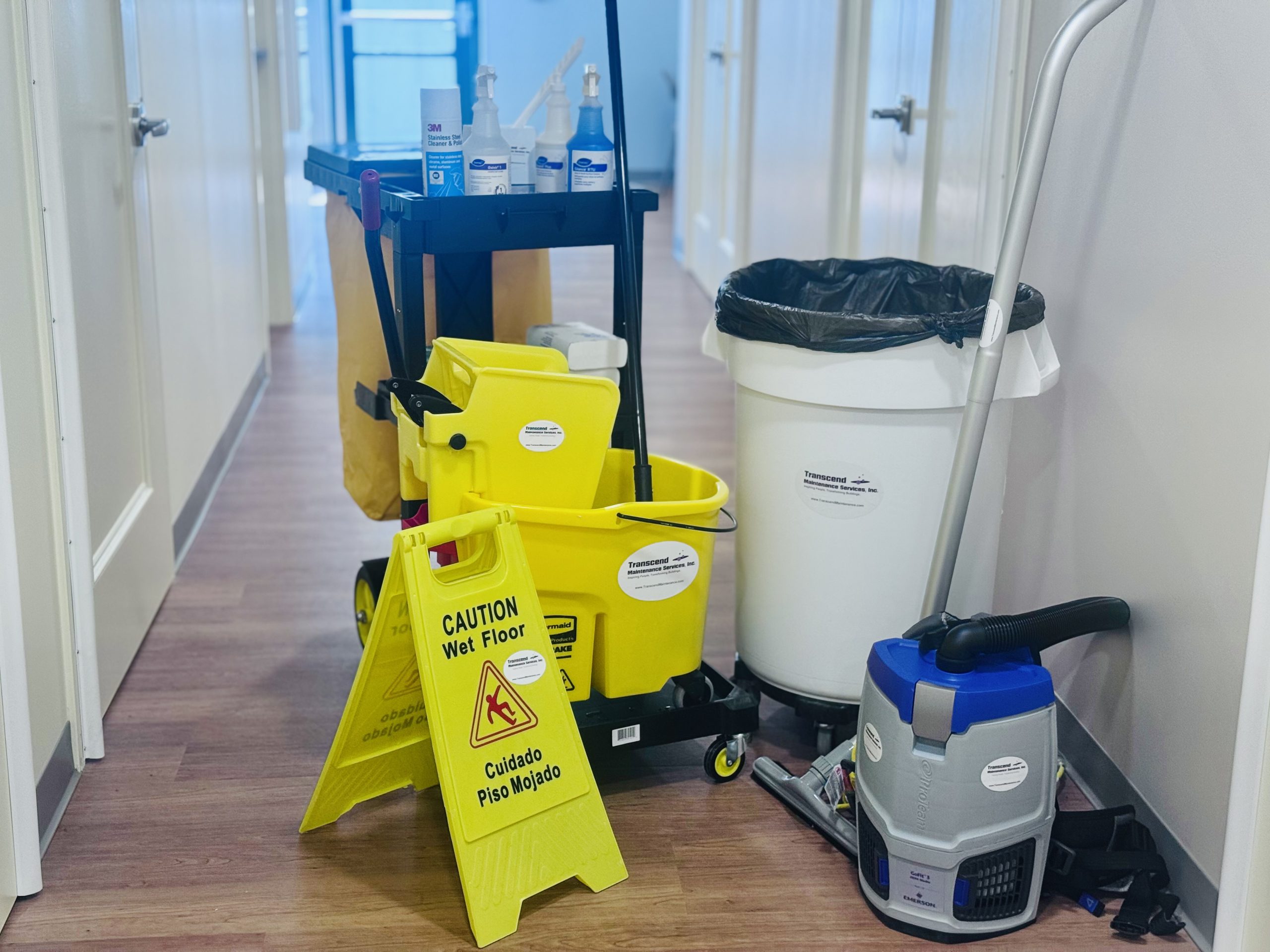 Janitorial cleaning cart with supplies, mop, and caution sign in office hallway, part of Transcend Maintenance commercial cleaning services.